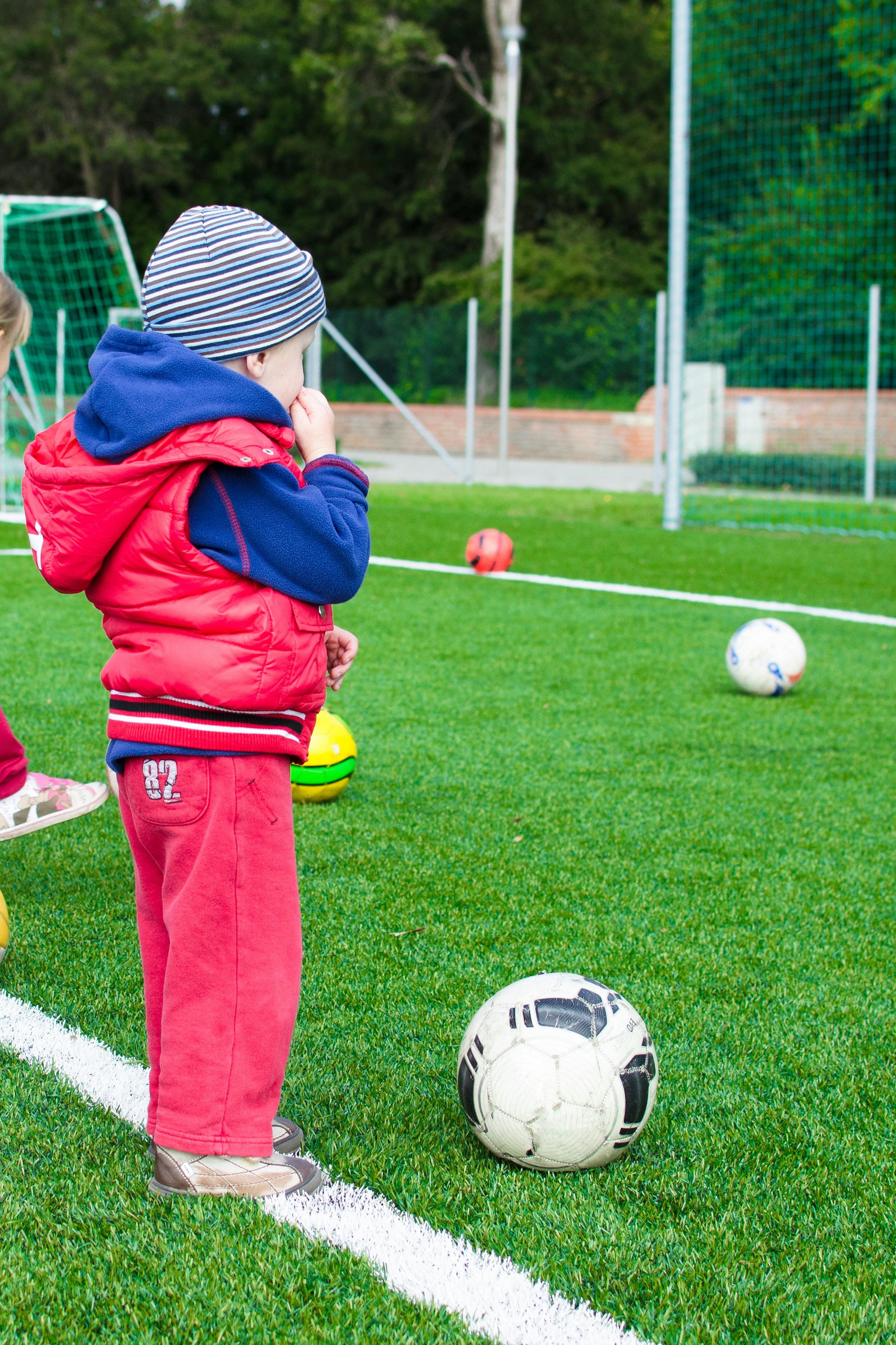 A toddler standing in front of a Soccer ball on a field.