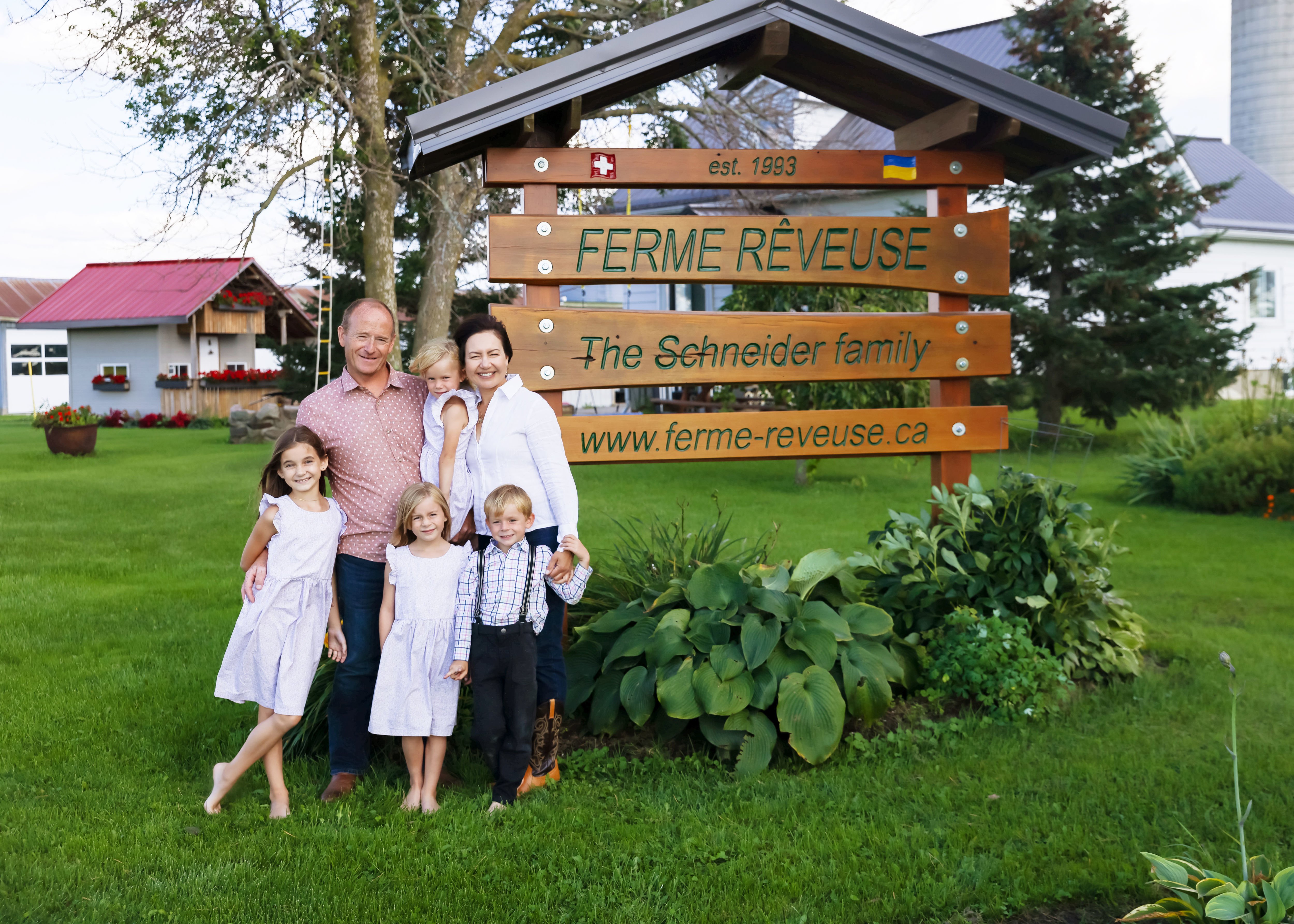 Les propriétaires Kornel et Olga Schneider et leurs quatre enfants devant un panneau en bois portant le nom de leur ferme, leur site web, leur histoire et leur origines.