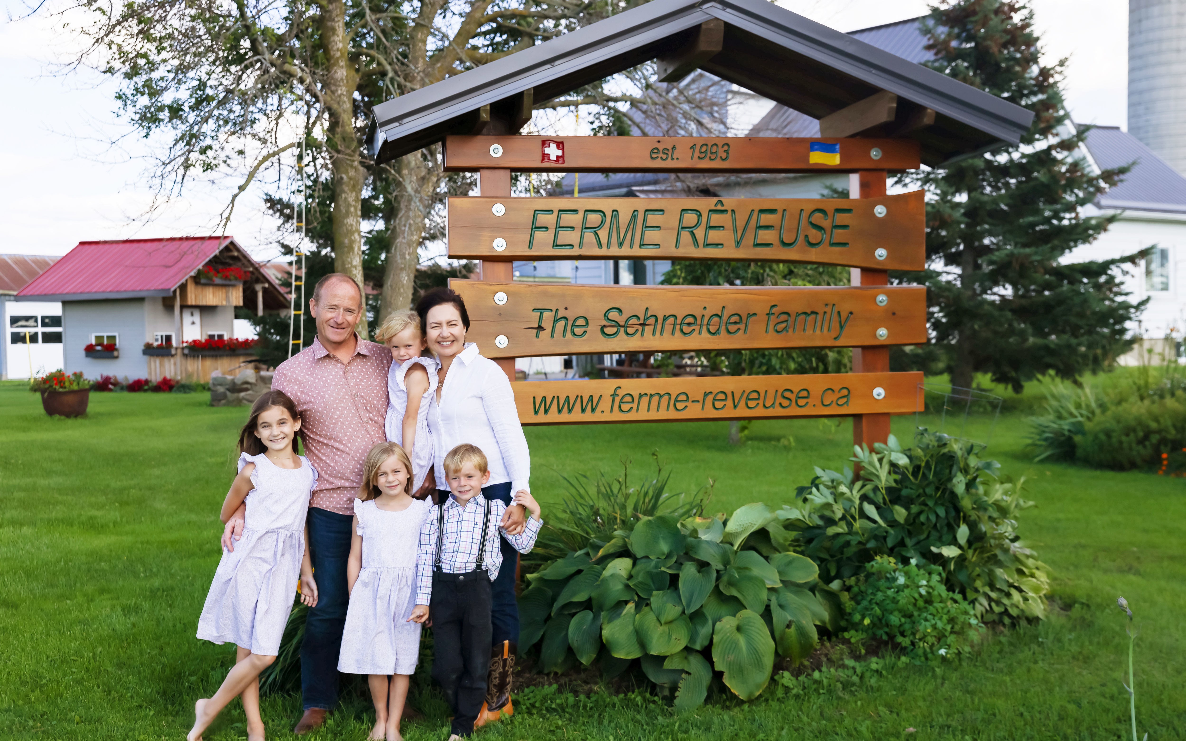 Les propriétaires Kornel et Olga Schneider et leurs quatre enfants devant un panneau en bois portant le nom de leur ferme, leur site web, leur histoire et leur origines.