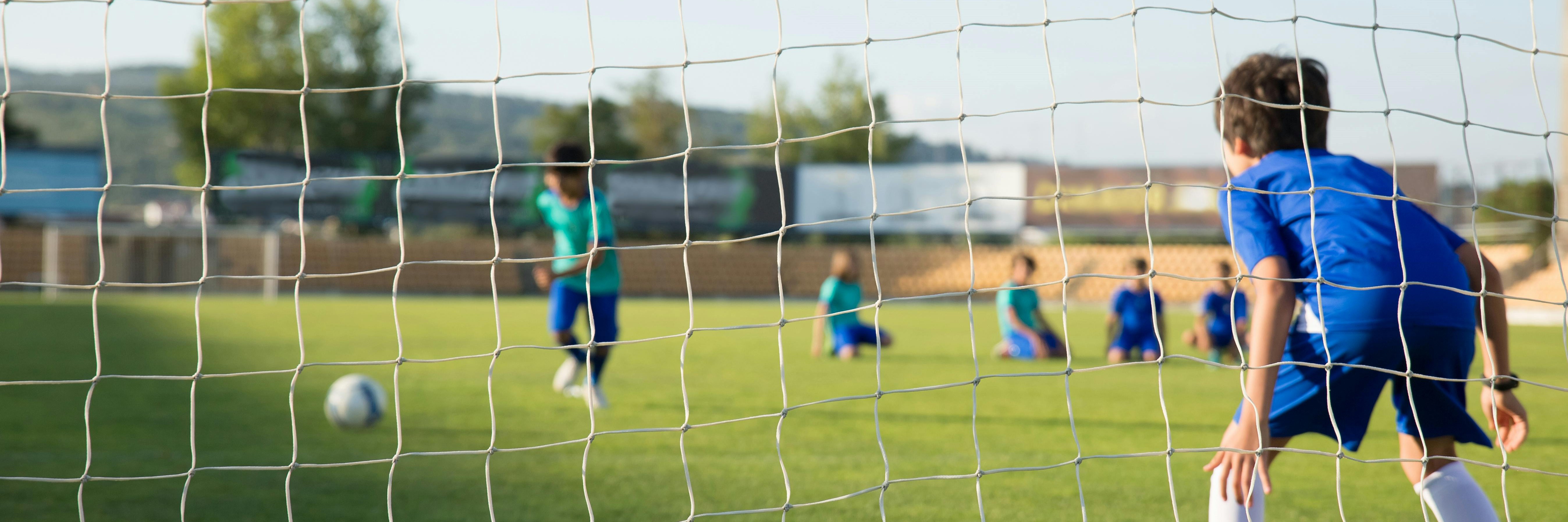 Kid in a soccer net looking at players with the soccer ball.