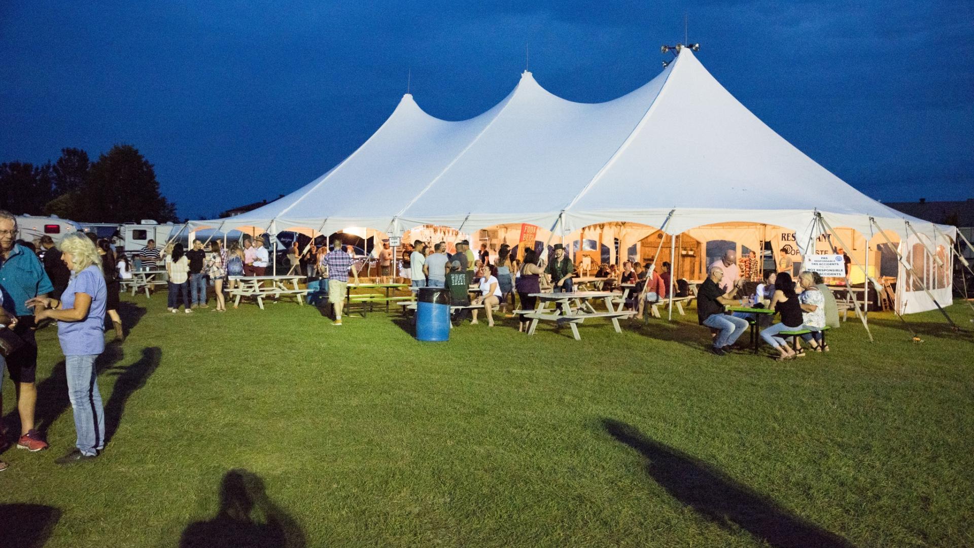 Large white tent with people gathered, set on a grassy field at dusk.