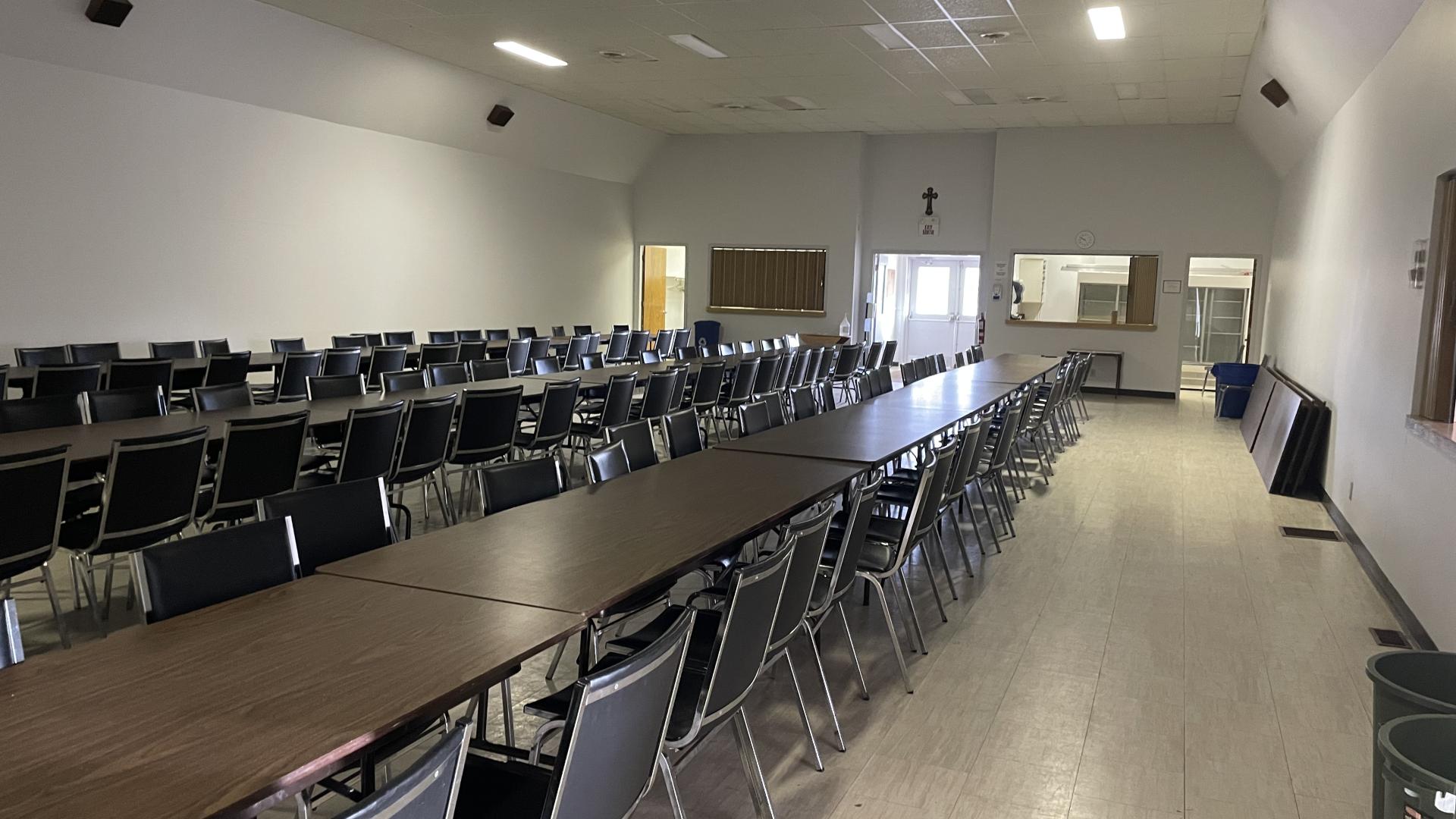 Empty meeting hall with rows of tables and chairs, white walls, and a high ceiling.