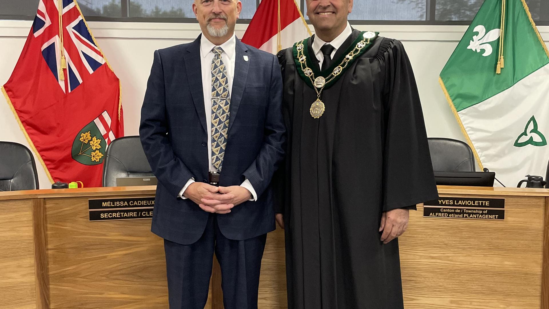Two men in a council chamber, one in a suit, the other in a ceremonial robe, smiling.