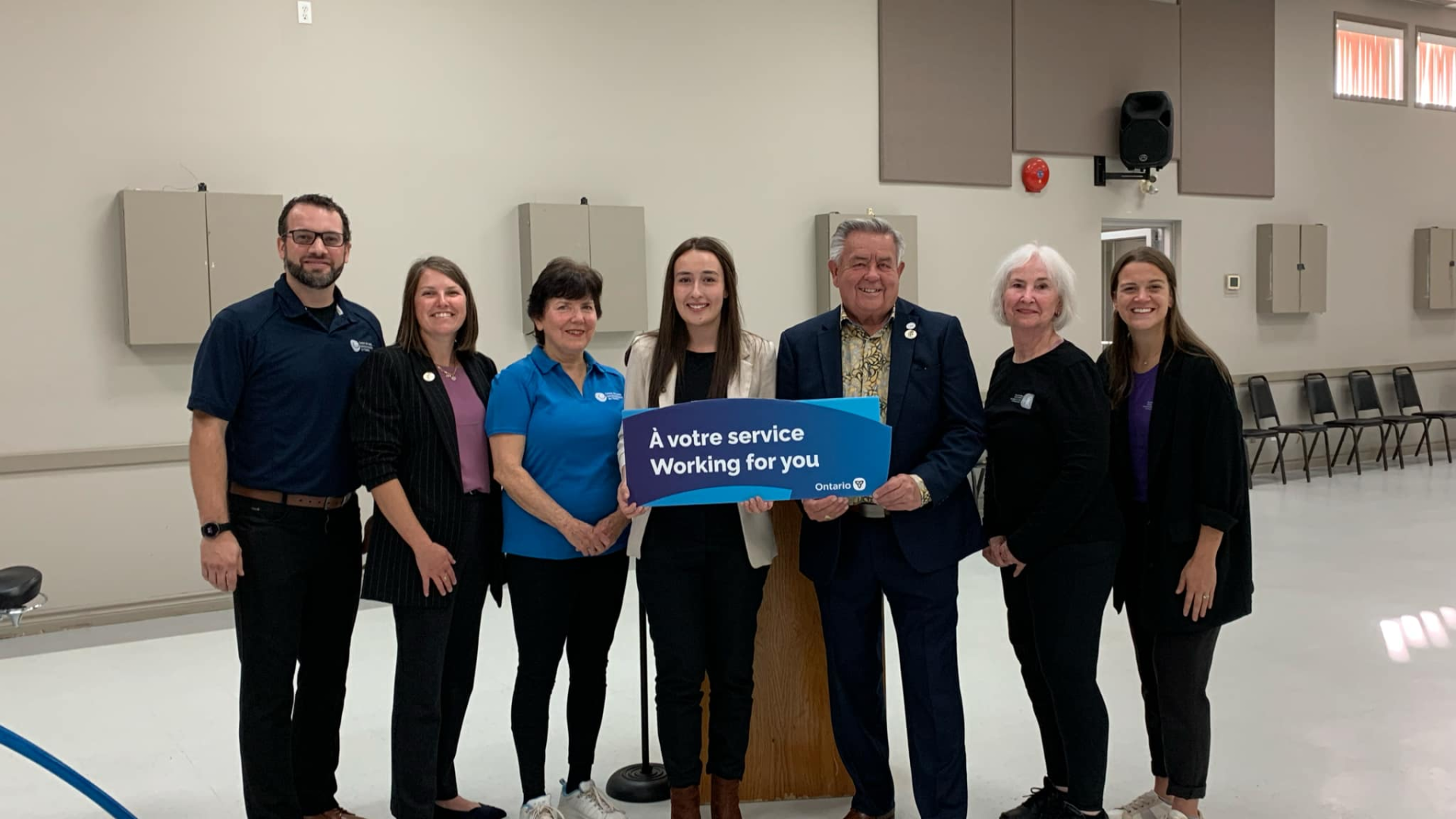 Seven people smiling in a hall, one holding a blue sign.