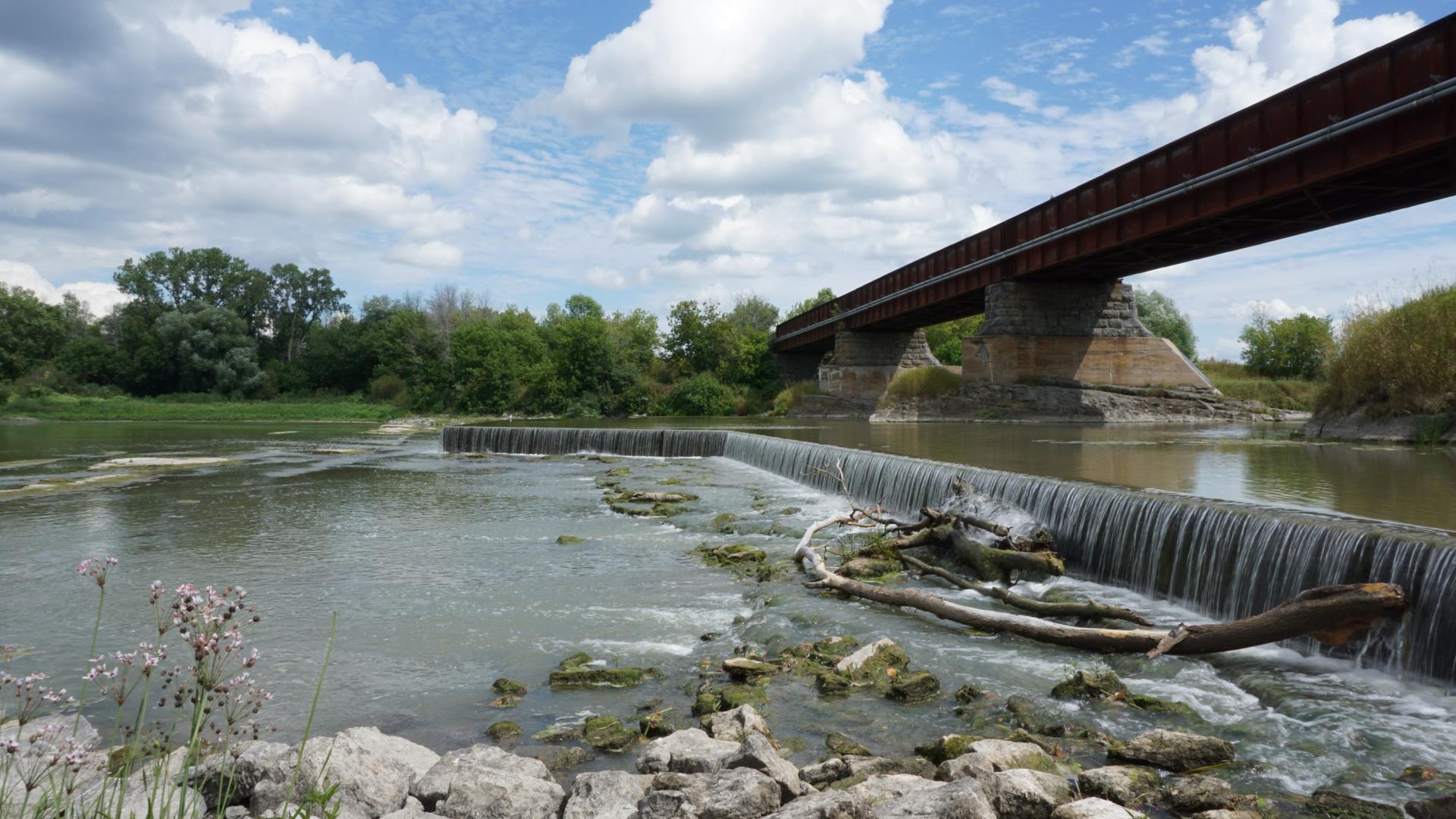 Pont en métal au-dessus d'une rivière avec cascade, ciel bleu et nuages.