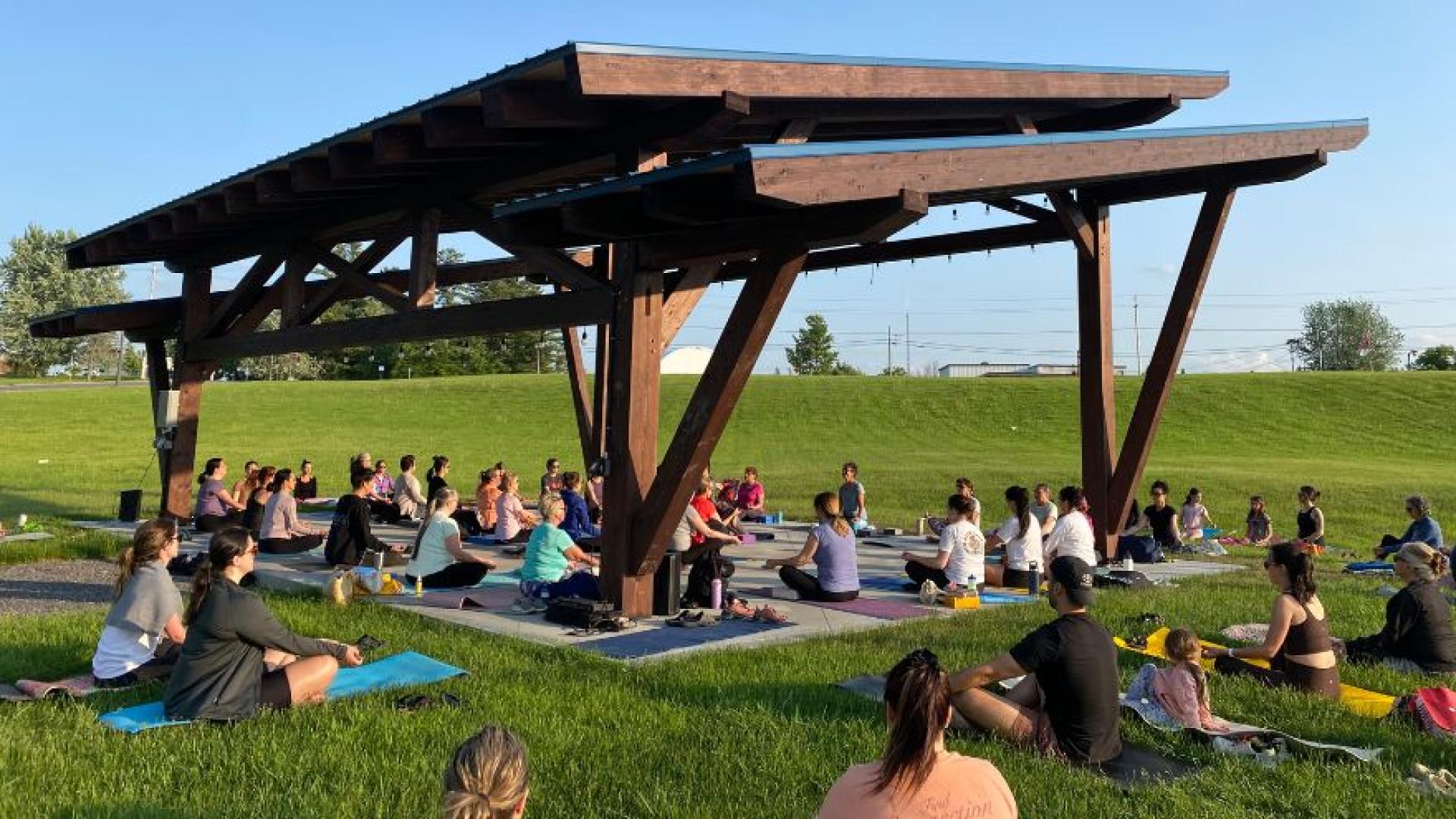 60 personnes assis au sol face à une instructrice de yoga sous une scène extérieure