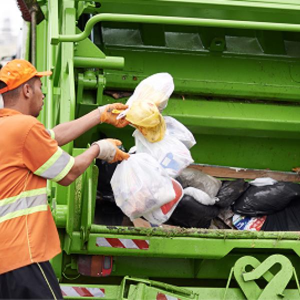 Sanitation worker in orange uniform loads trash bags into a green garbage truck.