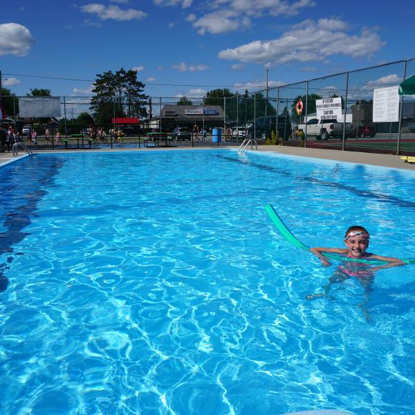 Child swimming in outdoor pool with a green float, under a clear blue sky.
