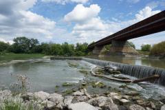 River with a small waterfall, bridge, and cloudy sky.